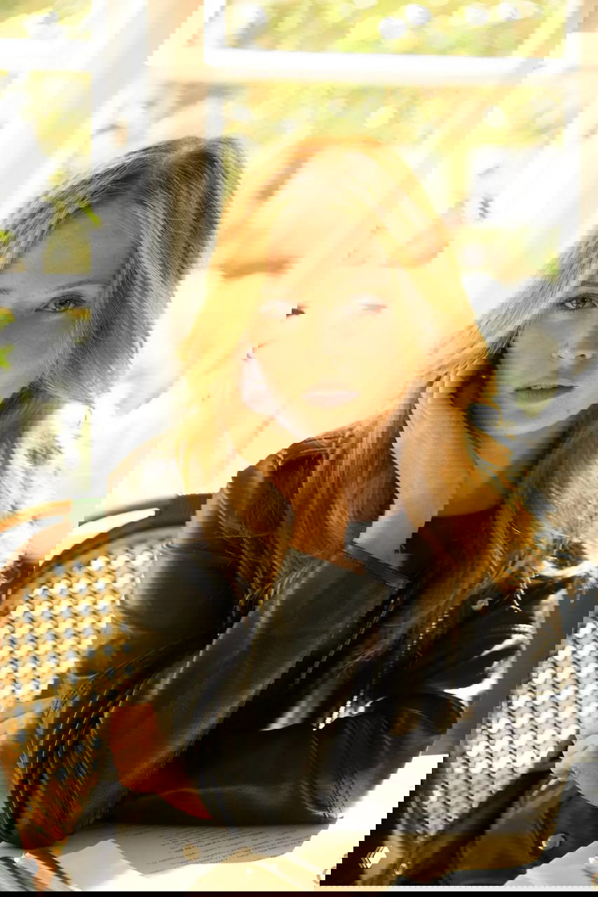 Крупный план женщины с распущенными светлыми волосами за столом в кожаной куртке, мягкий дневной свет и задумчивый взгляд.