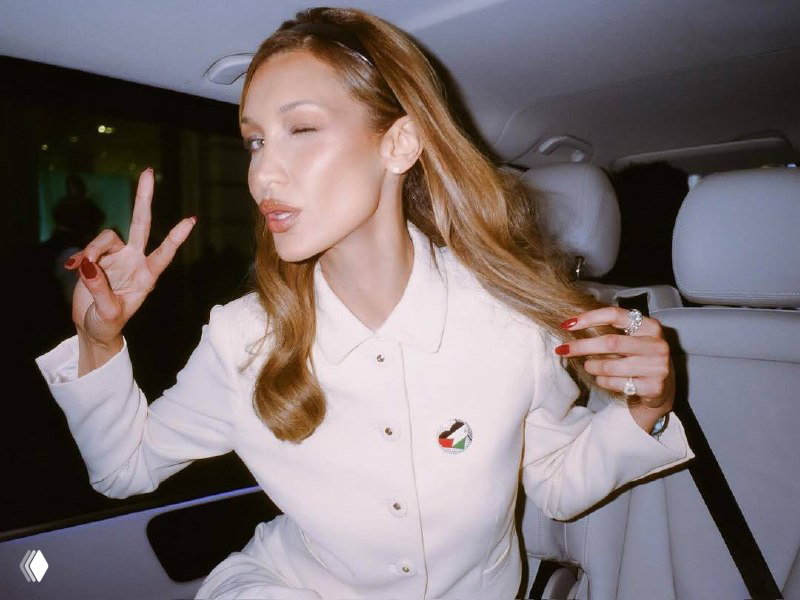 Белла Хадид в белом костюме в салоне автомобиля моргает и показывает жест V, заметны красный маникюр и украшения на пальцах.