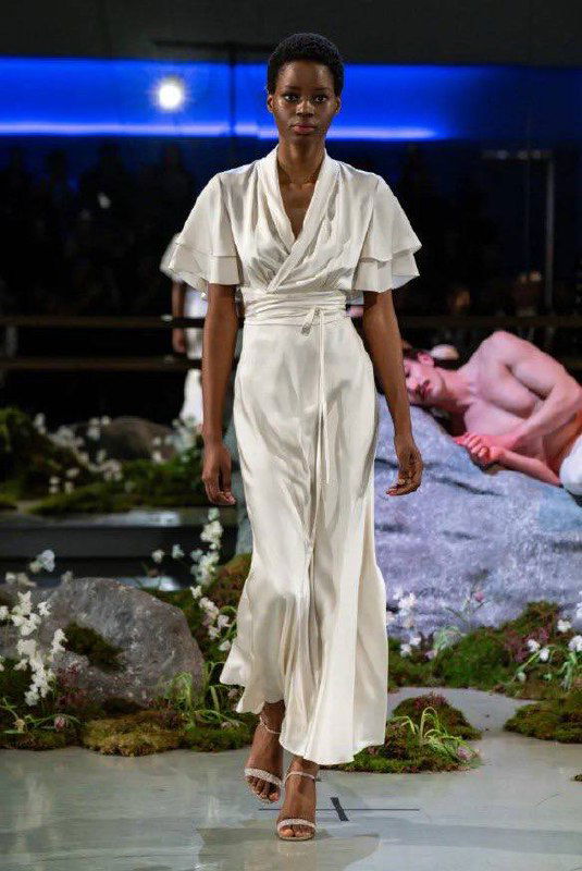 Модель в струящемся белом платье с воланами идёт по подиуму; в кадре видна художественная постановка шоу Патрика Макдауэлла.