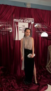 Женский образ на красной сцене Marie Claire: струящаяся блуза и длинная юбка, минималистичный аксессуар в руках, логотип мероприятия сзади.