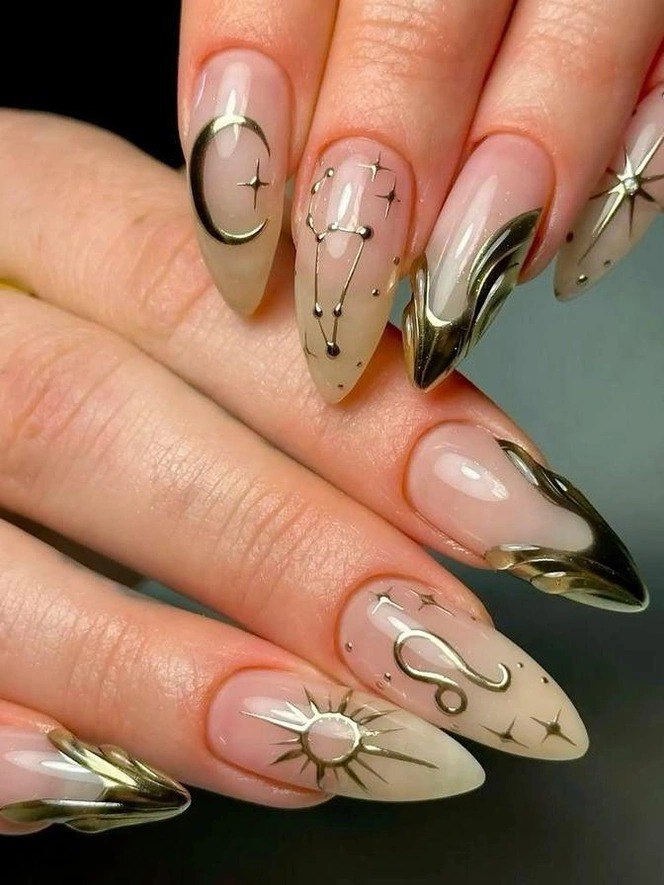 Острые миндалевидные ногти нюдового оттенка с золотыми металлическими символами — месяц, созвездие и тонкие акценты, аккуратный глянцевый финиш.