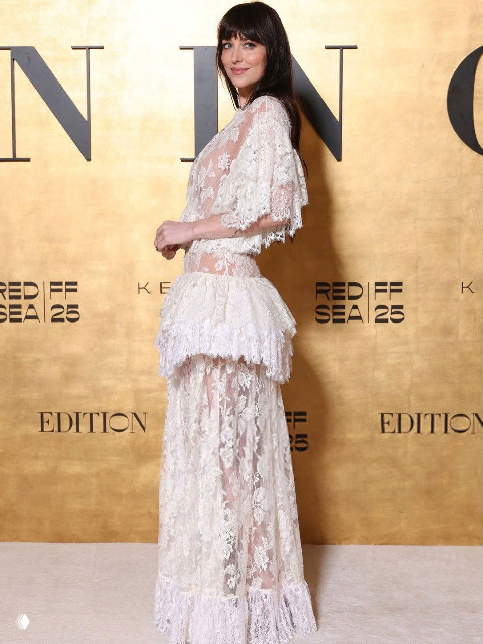 Профильный ракурс Дакоты Джонсон в кружевном наряде Chloé: вид сбоку, рука на бедре, волнистые волосы и золотая фотозона.