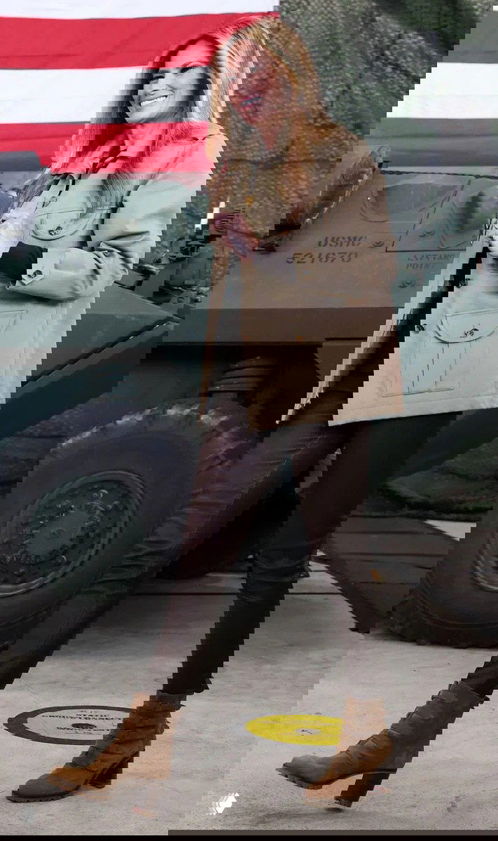 Женщина в бежевой куртке и коричневых кожаных брюках позирует на фоне военной техники и флага; образ с элементами утилитарного стиля.