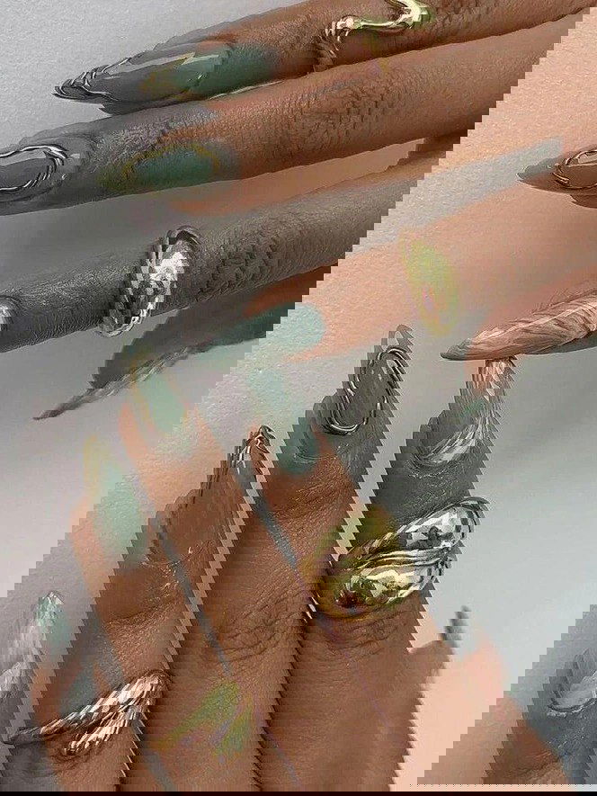 Мраморный зелёно‑мятный маникюр с золотыми объёмными рамками и глянцевым финишем на длинных ногтях, дополненный крупными золотыми кольцами на пальцах.