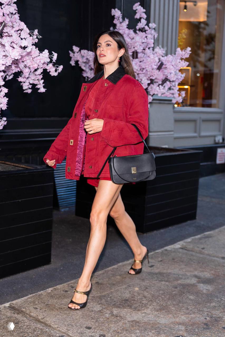 Моника Барбаро в кадре у цветущего дерева в алом total look: рабочая куртка, мини-юбка, чёрная сумка и тонкие босоножки.