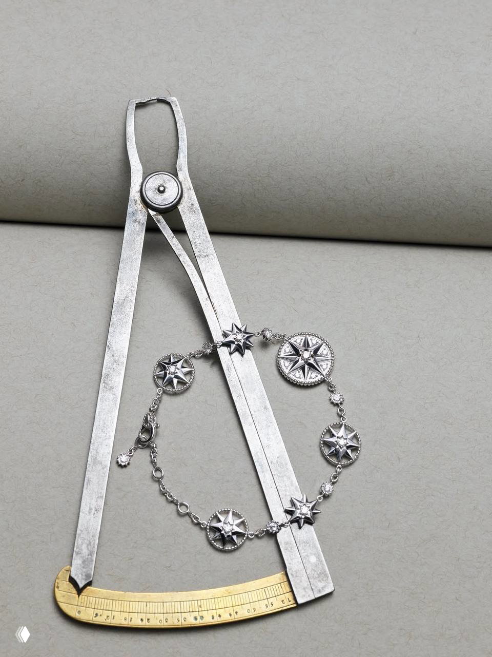 Серебряный браслет Dior с медальонами‑компасными розами выложен рядом с чертежным инструментом — художественный натюрморт рекламной кампании