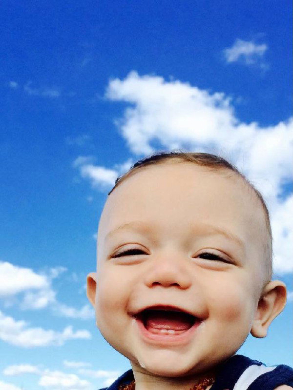 Крупный портрет маленького ребёнка с широкой улыбкой на фоне облачного неба — радостный момент из семейной подборки снимков.