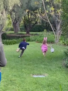 Динамичный кадр: дети бегут по газону с пасхальными корзинками в руках; охота за яйцами на свежем воздухе среди зелёных деревьев.