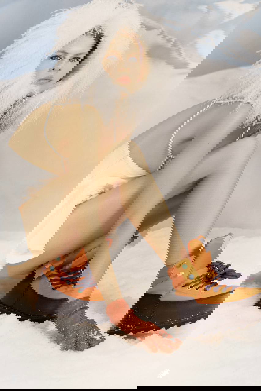 Модель в белой перьевой шапке и лёгком мини‑наряде сидит на снегу — сочетание хрупкой красоты, прозрачности и контрастной обуви в 90‑х стиле.