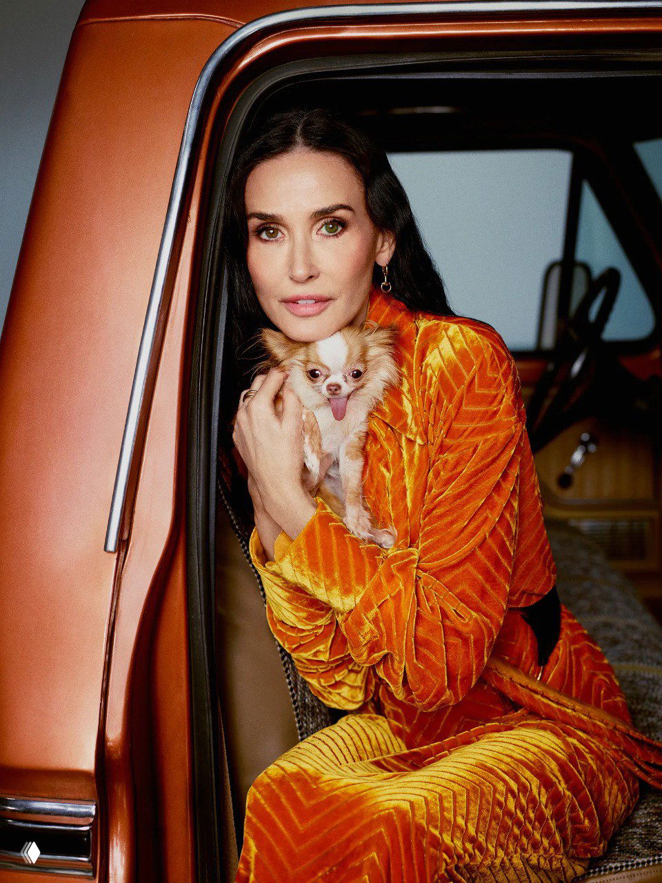 Женщина в ярком бархатном оранжевом костюме держит маленькую собачку в руках у двери ретро‑автомобиля в студийной постановке.