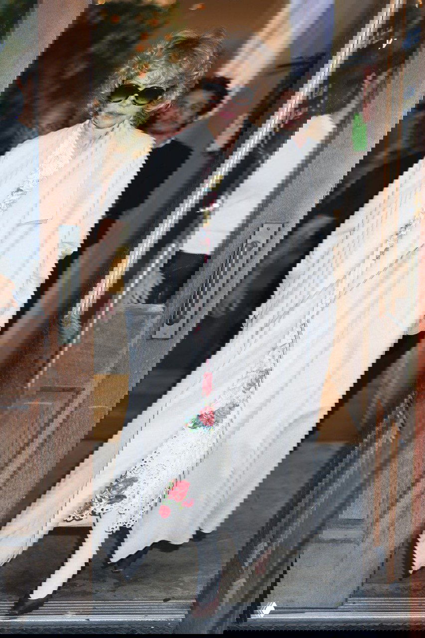 Памела Андерсон выходит из дверей pop-up пространства: длинное пальто поверх цветочного платья, солнцезащитные очки и сотрудники мероприятия рядом.