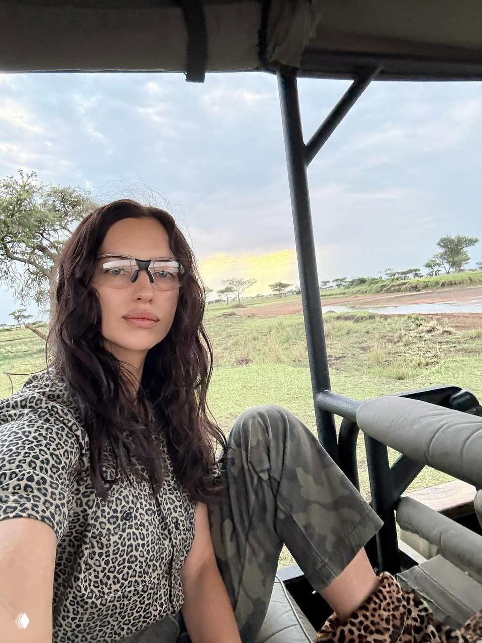 Селфи в сафари-джипе: женщина в камуфляжных брюках и леопардовой обуви на фоне саванны и низкого неба перед закатом.