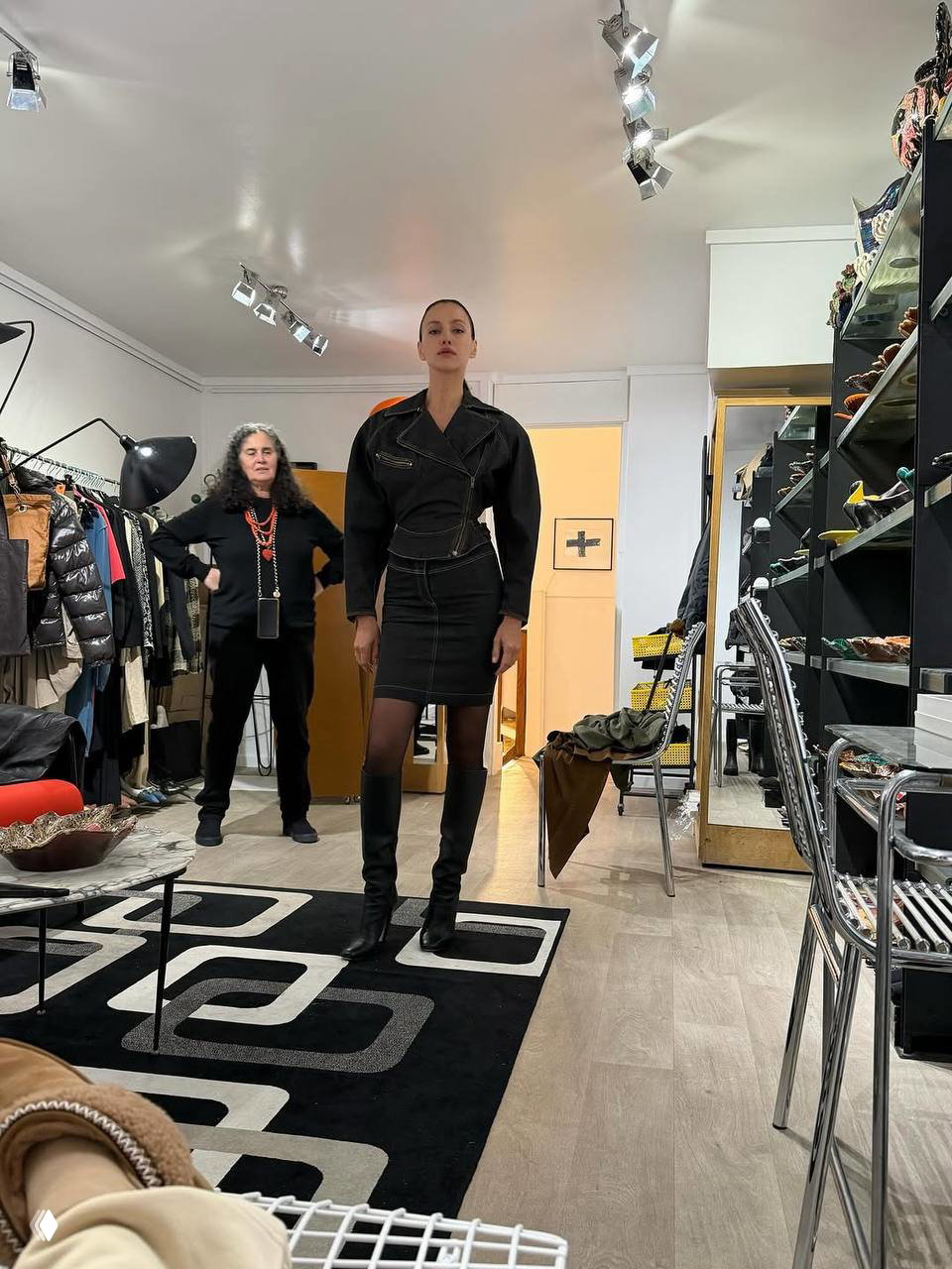 Примерка в бутике: модель в тёмном платье и высоких сапогах позирует в окружении одежды, обуви и зеркал в интерьере шоурума.