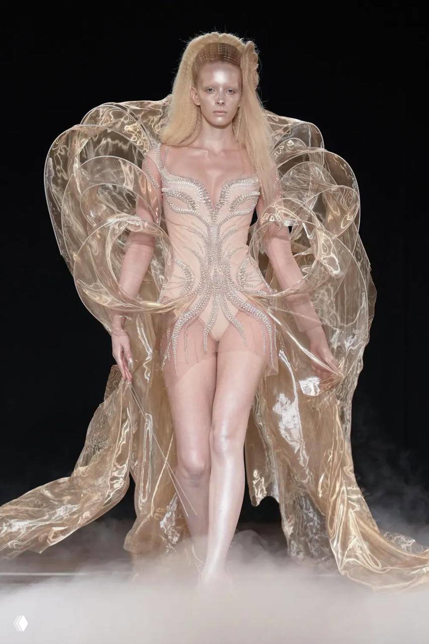 Модель в полупрозрачном скульптурном платье Iris Van Herpen на подиуме Недели высокой моды в Париже, окружена дымом и театральным светом.