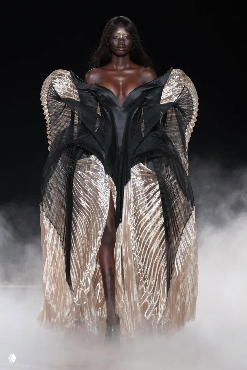 Темнокожая модель в объёмном платье с крыльями Iris Van Herpen; текстурные складки, драматичный контраст и туманная сцена модного показа.