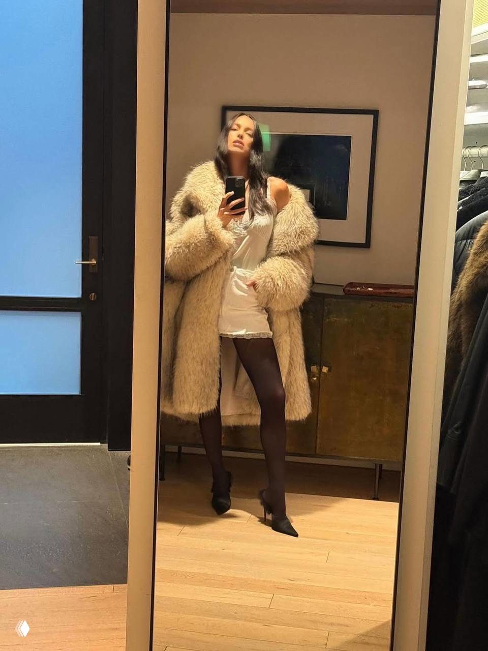 Отражение в зеркале: женщина в пушистом меховом пальто и мини-платье делает селфи в гардеробной на фоне картины и комода.