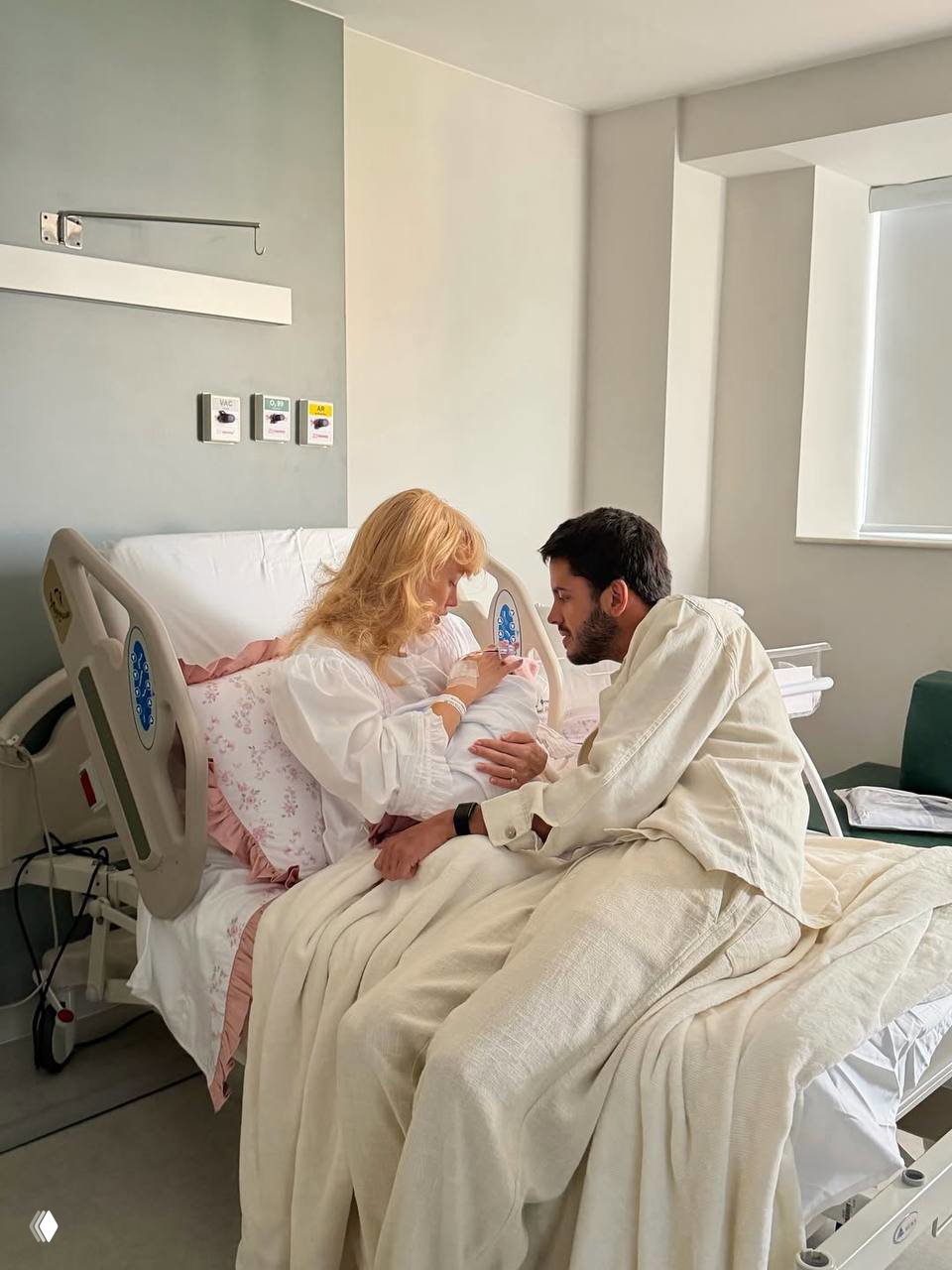 Мама в больничной палате держит новорождённую на руках, рядом отец наклонился и смотрит на малышку — трогательный семейный кадр.