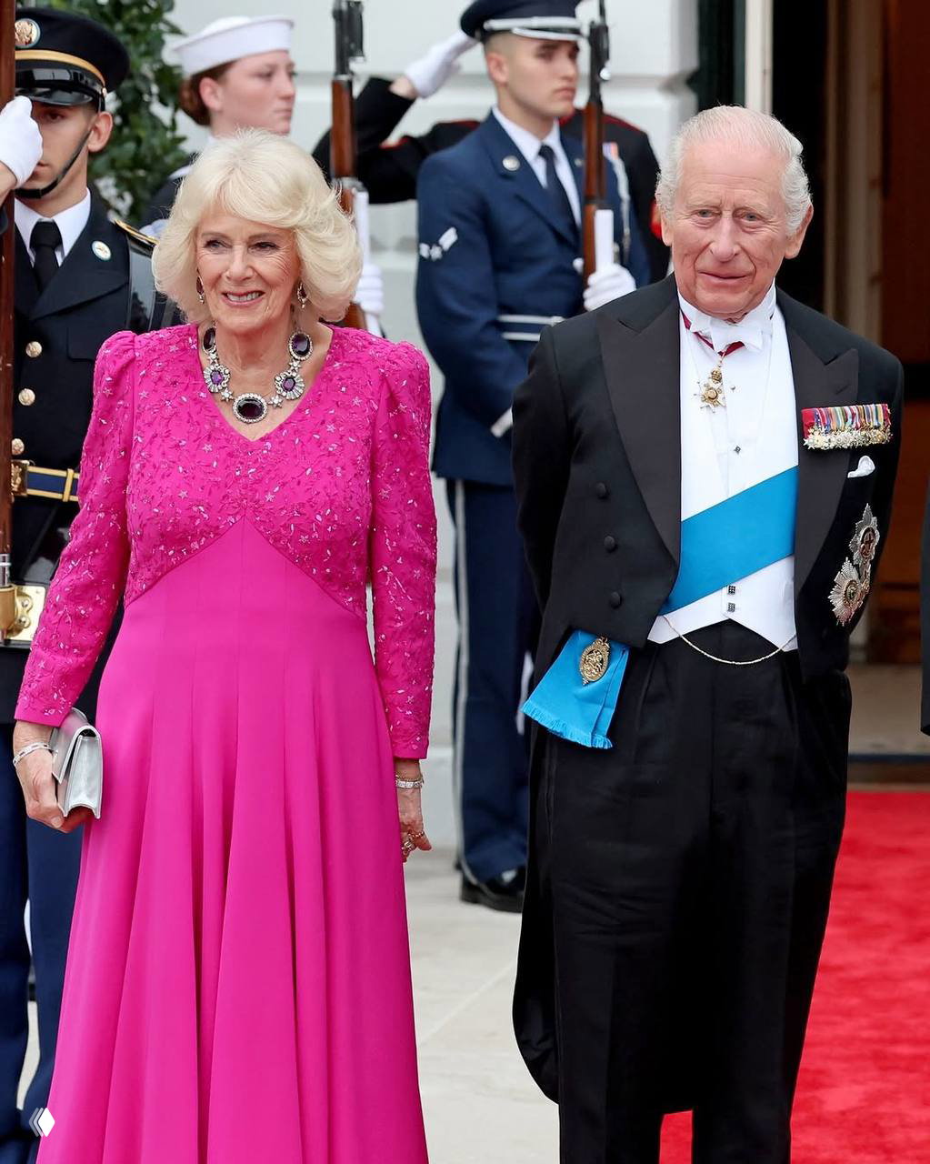 Пара в форме и женщина в ярком розовом платье позируют у здания, рядом видны караульные с винтовками.