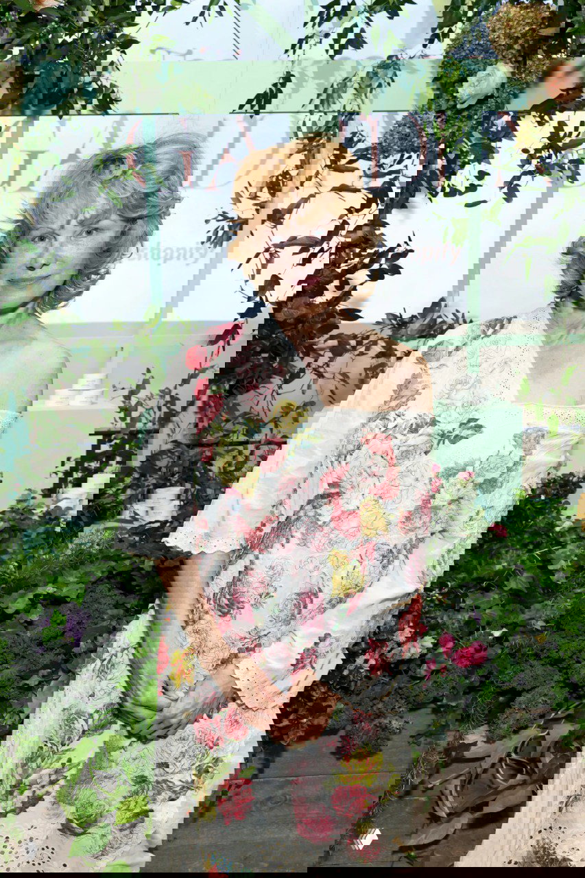 Портрет Памелы Андерсон на открытии: натуральный макияж и лёгкая улыбка, платье с розовым цветочным принтом на фоне зелёной растительности и декора.