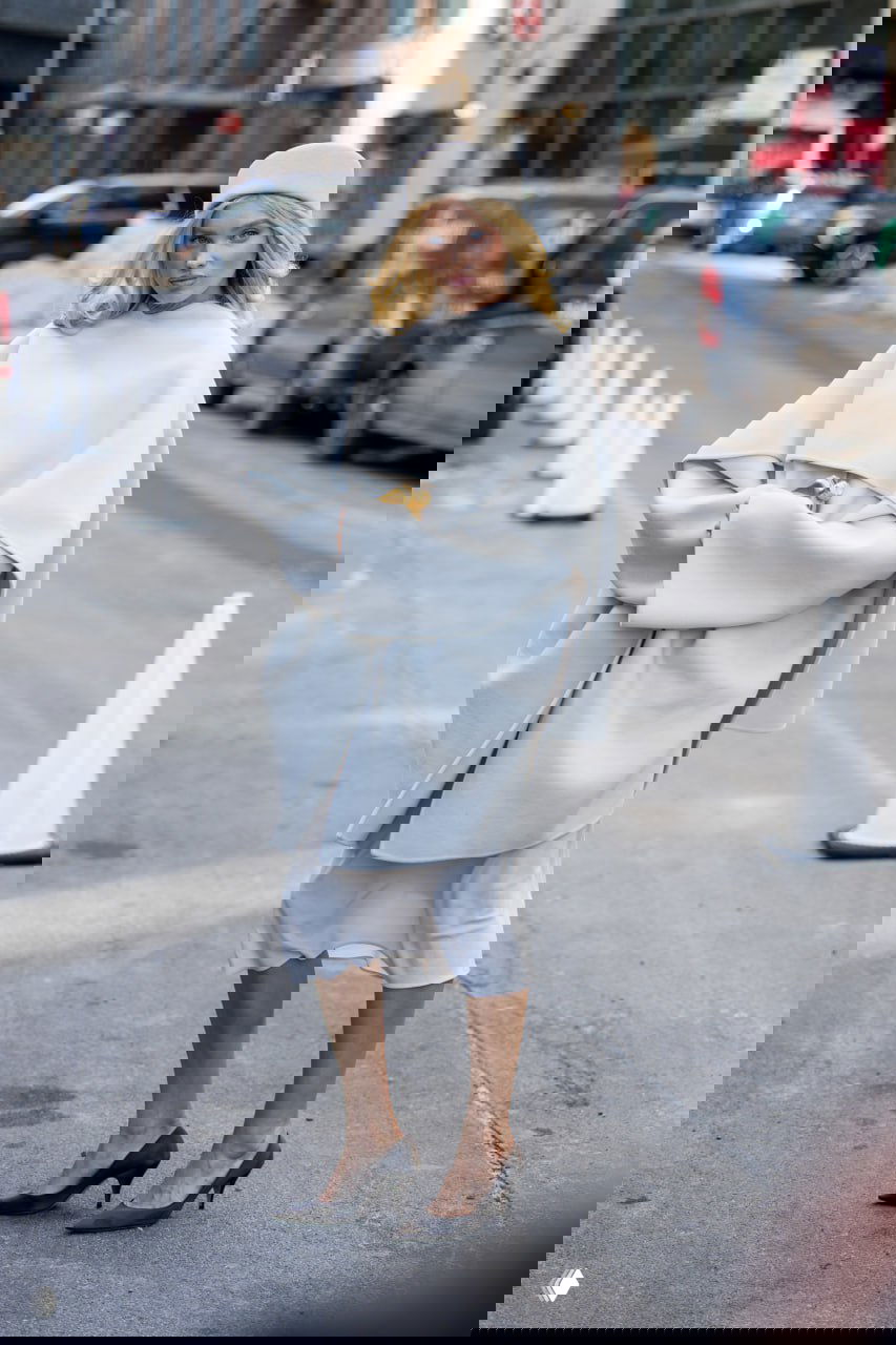 Женщина в светлом объёме‑пальто и шапке идёт по улице; на фоне автомобили и городская сцена в Нью‑Йорке.