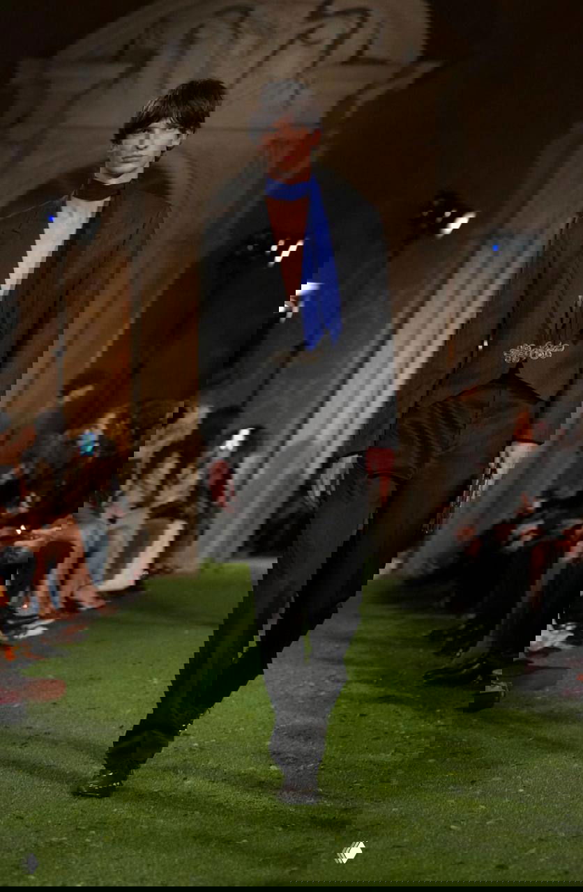 Модель в тёмном пиджаке с глубоким декольте и синий шарф на фоне архитектуры зала во время показa Valentino в Риме.