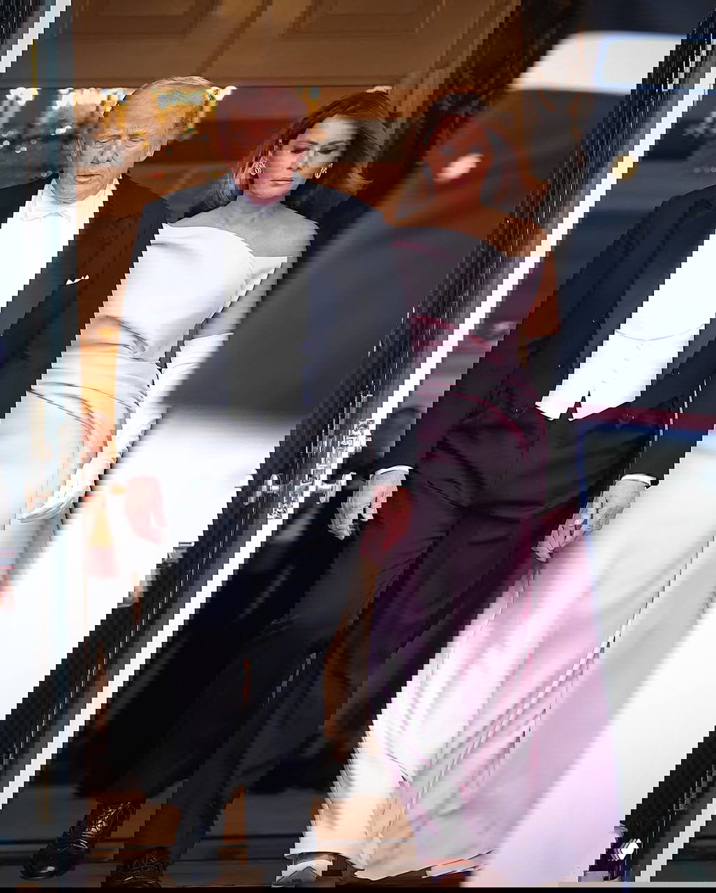 Мужчина в фраке и женщина в бледно-розовом вечернем платье выходят из здания Белого дома во время торжественного события.