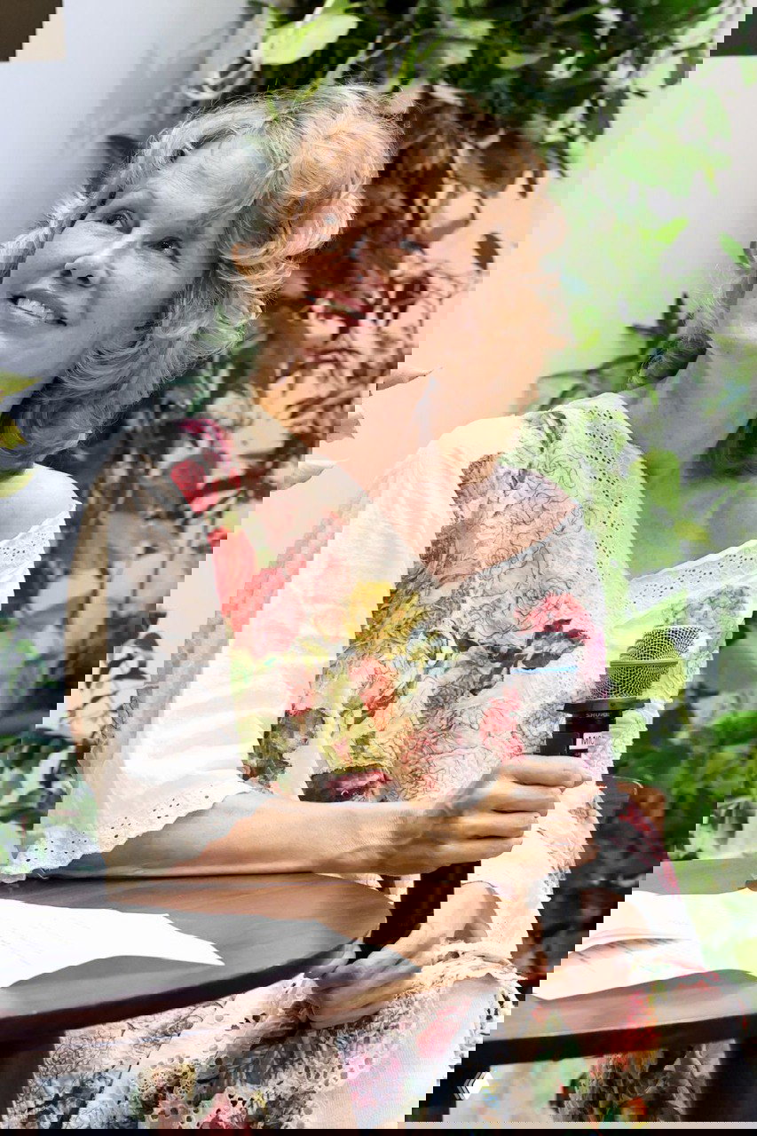 Памела Андерсон во время выступления с микрофоном в руках, в платье с крупным цветочным принтом на фоне зелёных растений и брендового оформления.