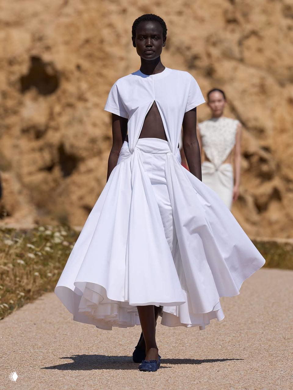 Показ Jacquemus Le Bonheur на Корсике: модель в белом платье‑рубашке с выразительными вырезами и пышной юбкой на фоне пейзажа.