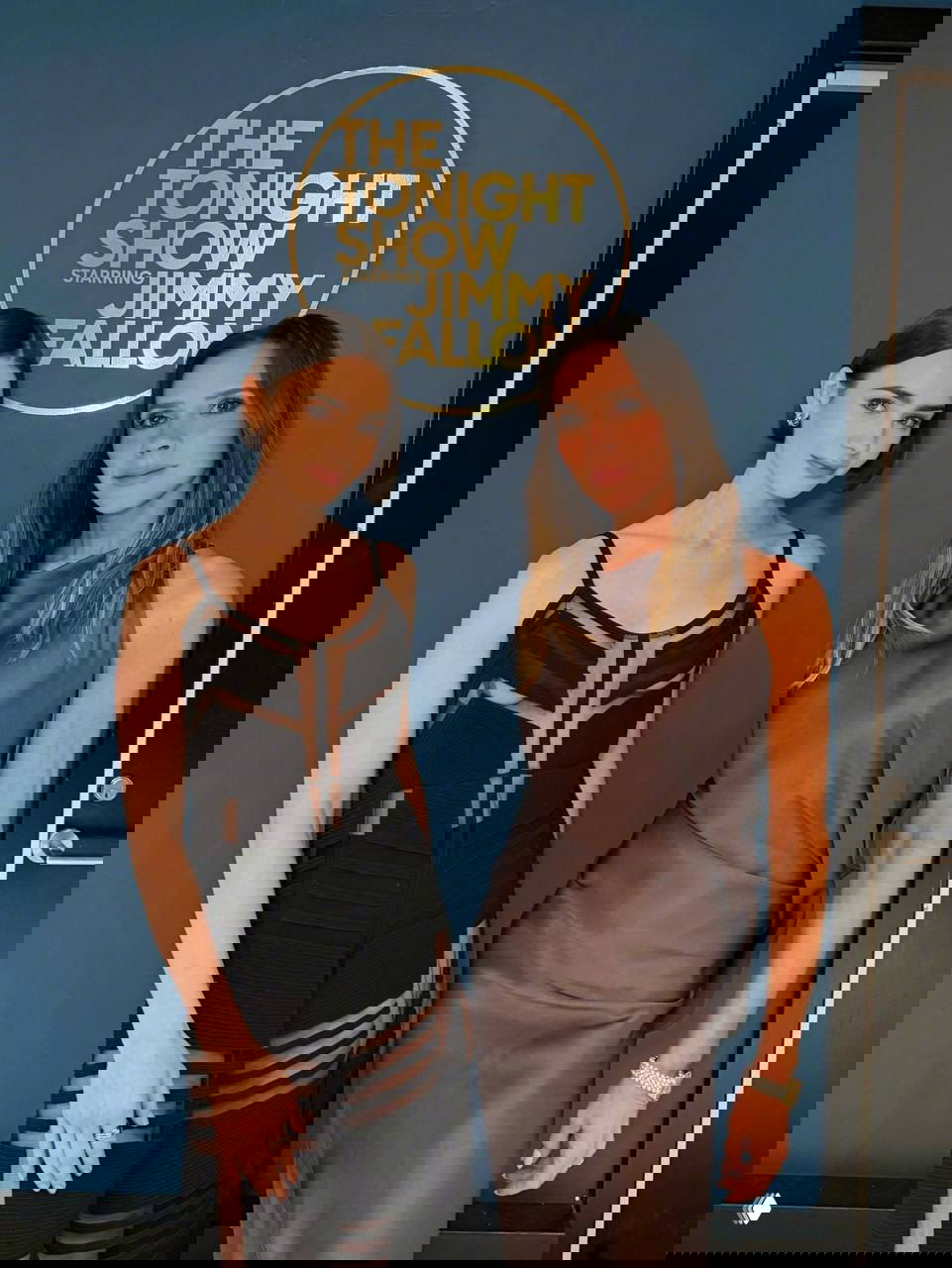 Две женщины позируют вместе у фона с логотипом телешоу, одна в чёрном прозрачном платье и ботфортах, другая в тёмном шёлковом платье и босоножках