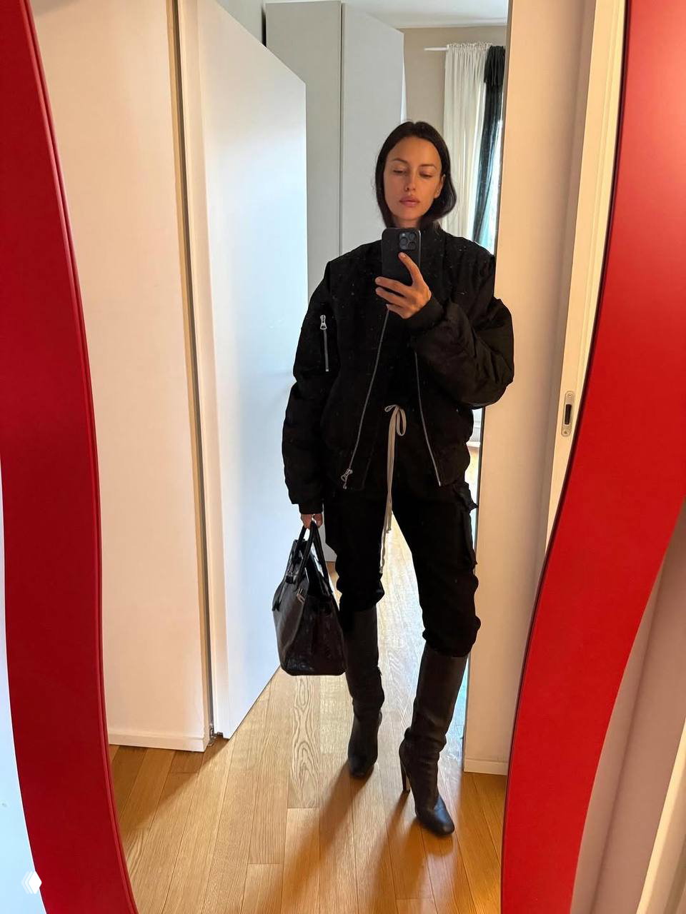 Полный рост у зеркала: женщина в чёрной тёплой куртке, с сумкой и сапогами делает снимок перед выходом, интерьер прихожей с деревянным полом.