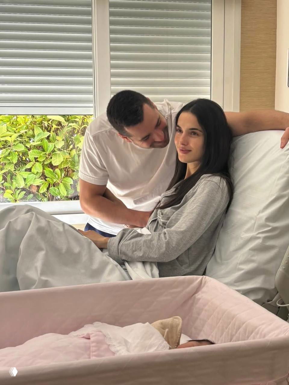 Бланка Падилья лежит в палате рядом с кроваткой, муж стоит рядом и поддерживает её, семейная сцена в больничной комнате.