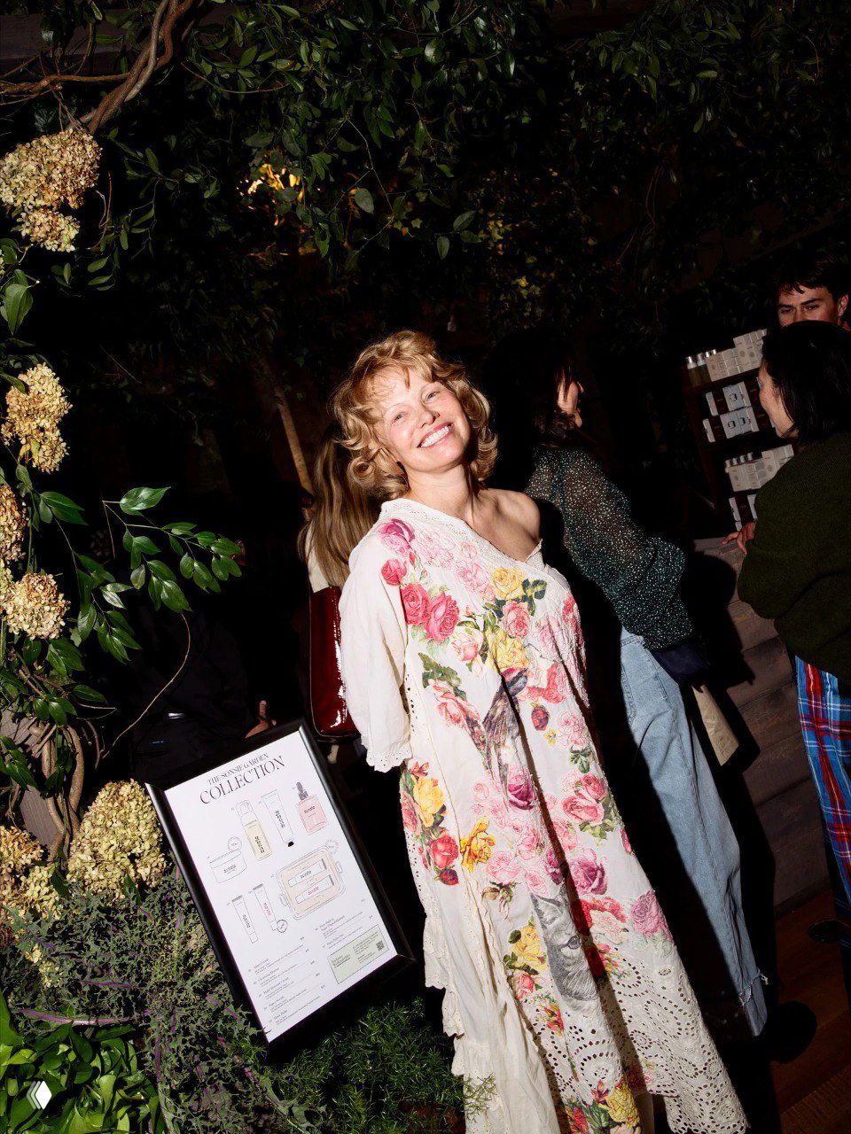 Памела Андерсон улыбается внутри pop-up пространства среди растений и гостей в свободном платье с разноцветными цветочными мотивами и декоративной стойкой с продукцией.