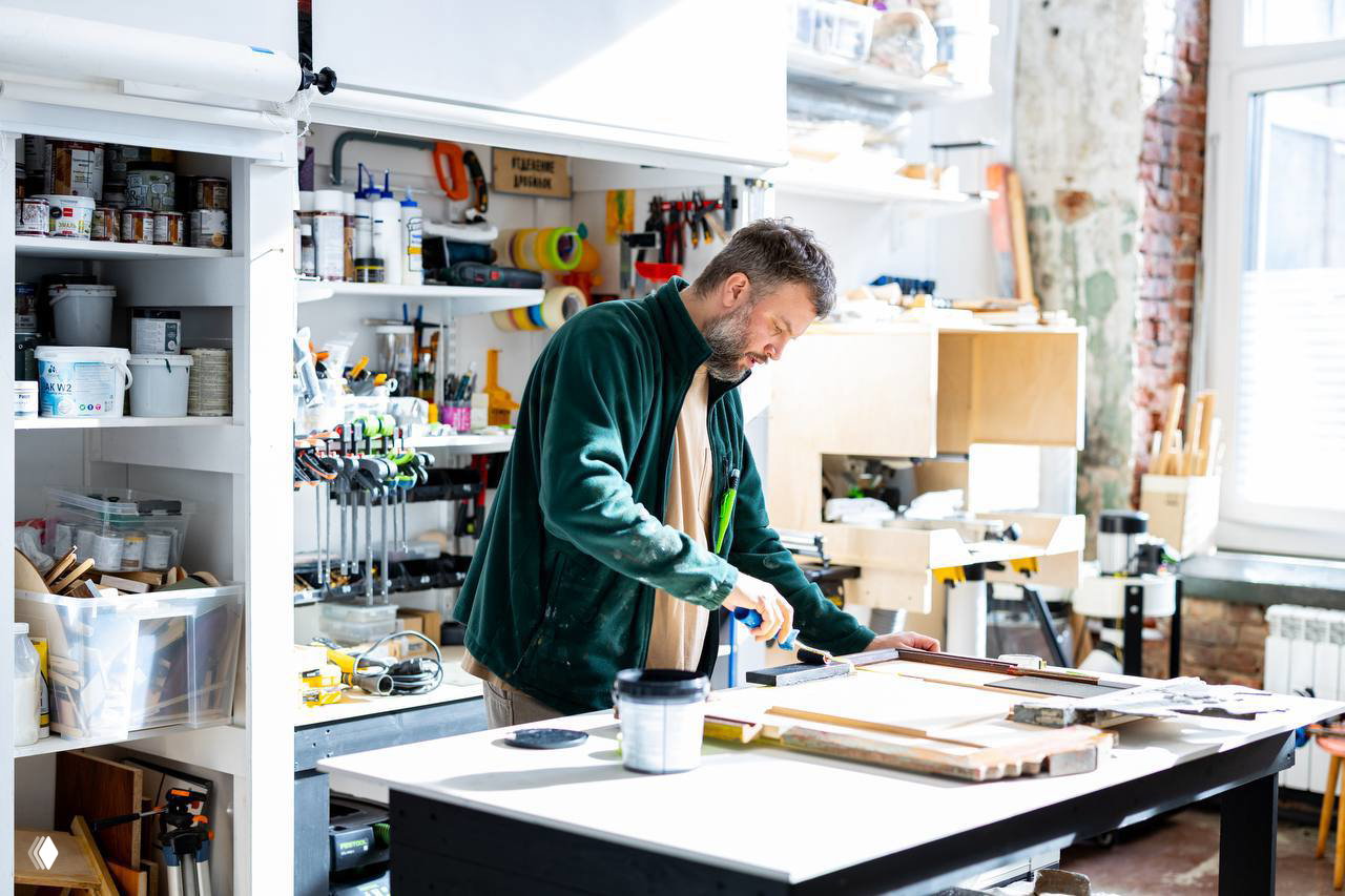 Художник в мастерской за рабочим столом с инструментами и материалами готовит инсталляцию для выставки: клеи, краски и заготовки на фоне кладовой и окон.