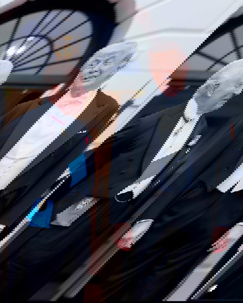 Два мужчины в парадной униформе и вечернем костюме стоят рядом на церемонии, виден обмен взглядами на крыльце.