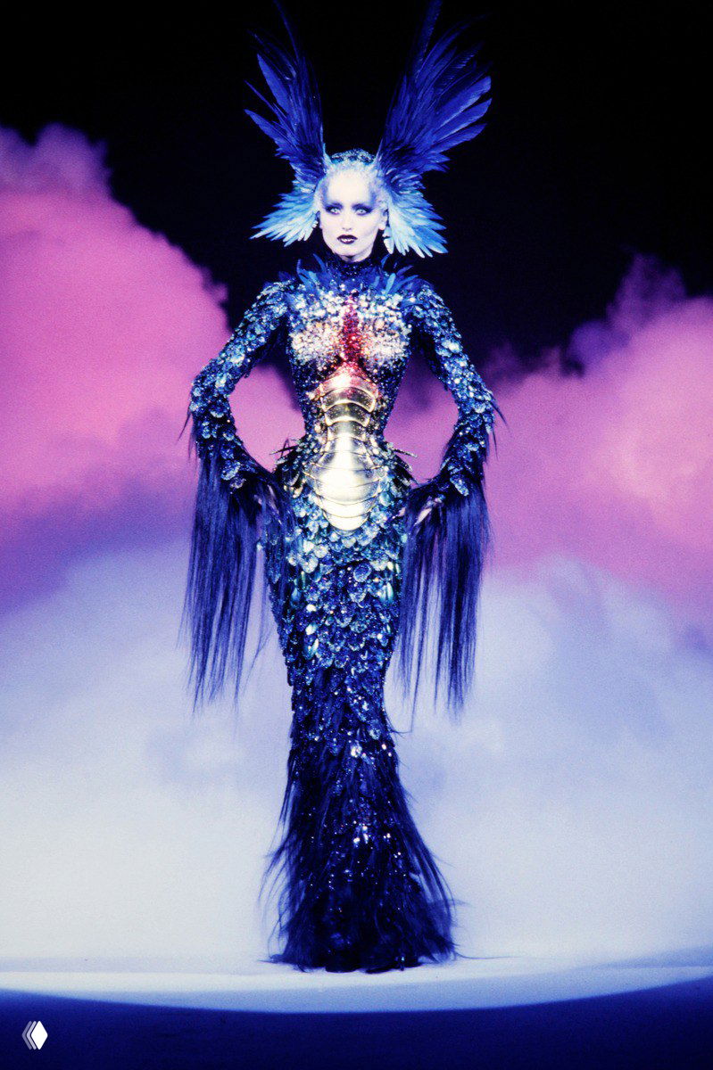 Цветной сценический образ: модель в перьевой и чешуйчатой композиции на фоне дымовой подсветки — эффектный театральный костюм.