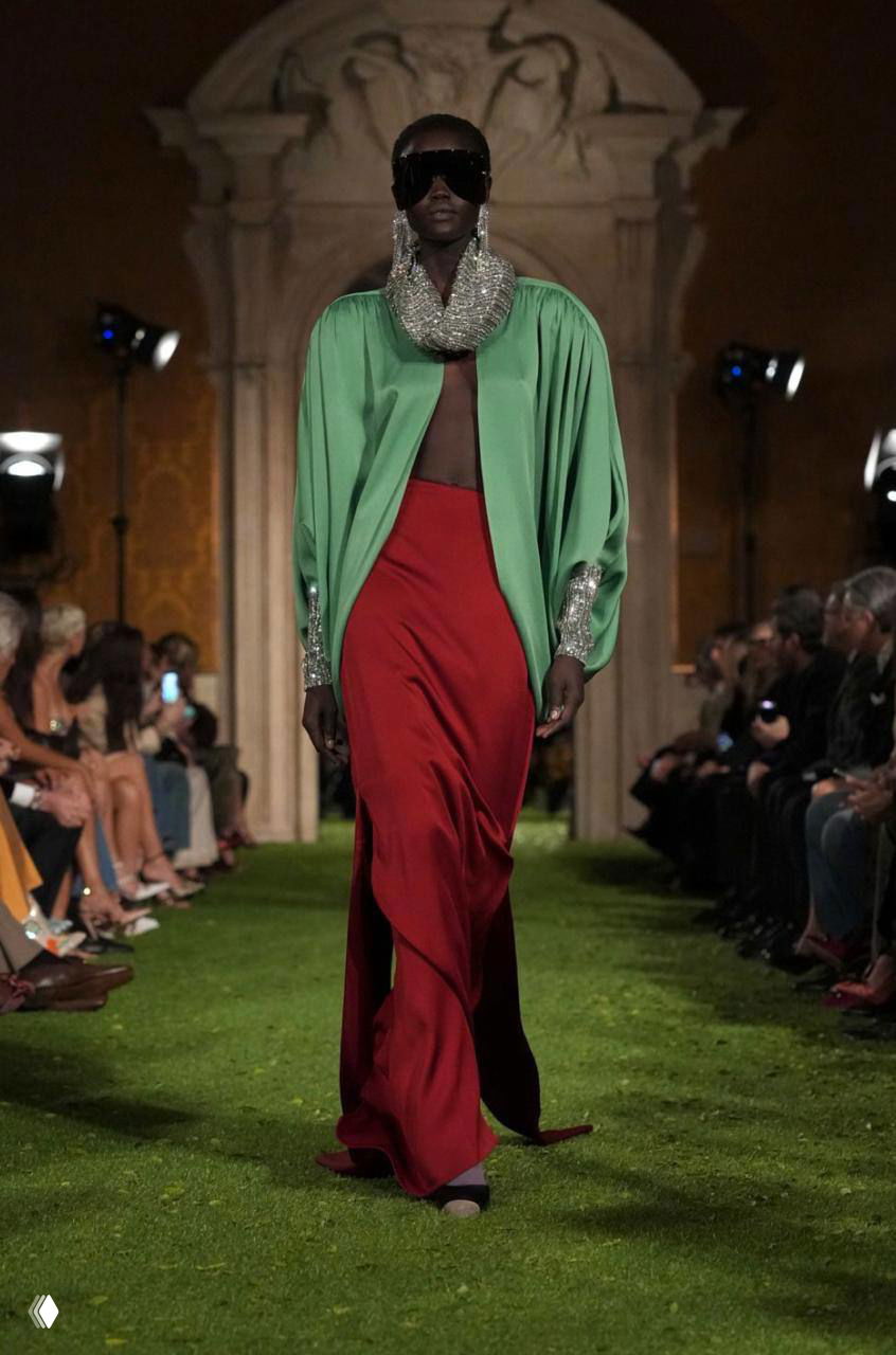 Модель в длиннейшем красном и зелёном образе с крупным украшением на шее дефилирует по травяному подиуму Valentino в Риме.