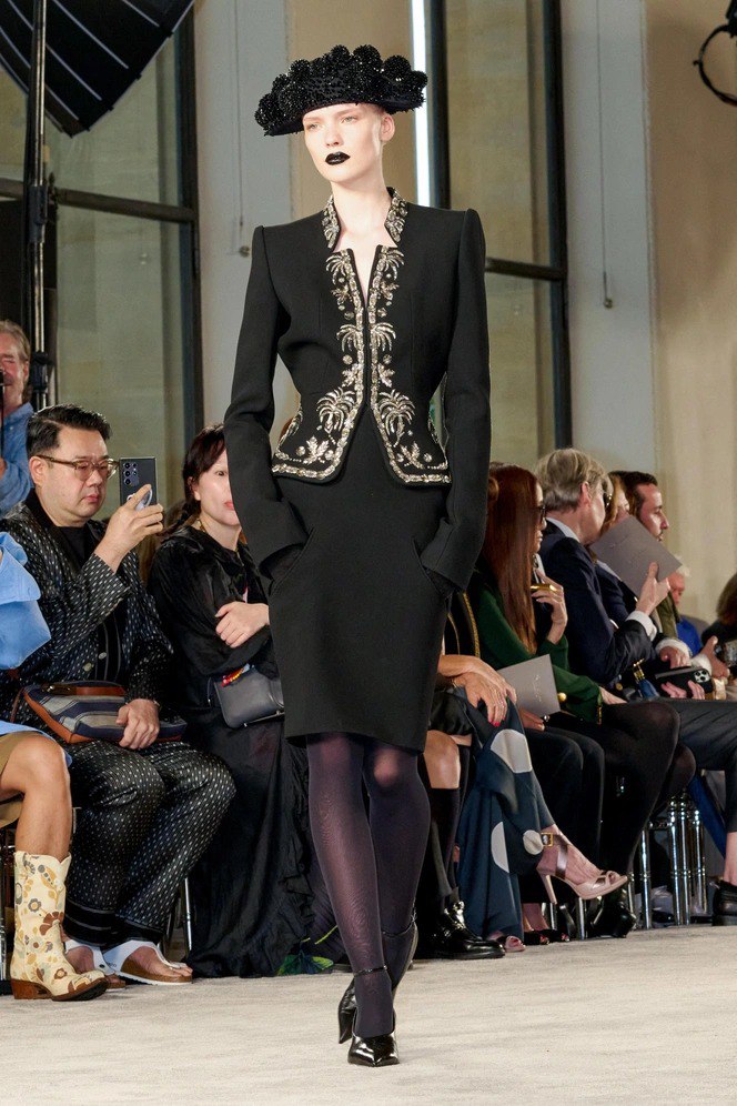 Модель в строгом чёрном костюме на подиуме, губы покрыты тёмной готической помадой; фоторепортаж с показа показывает контраст классики и вамп‑эстетики
