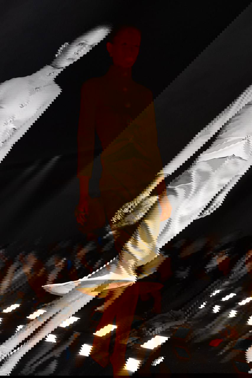 Модель в светлом платье и тонкой талии шагает по освещенному подиуму, кадр с длинной выдержкой на показе