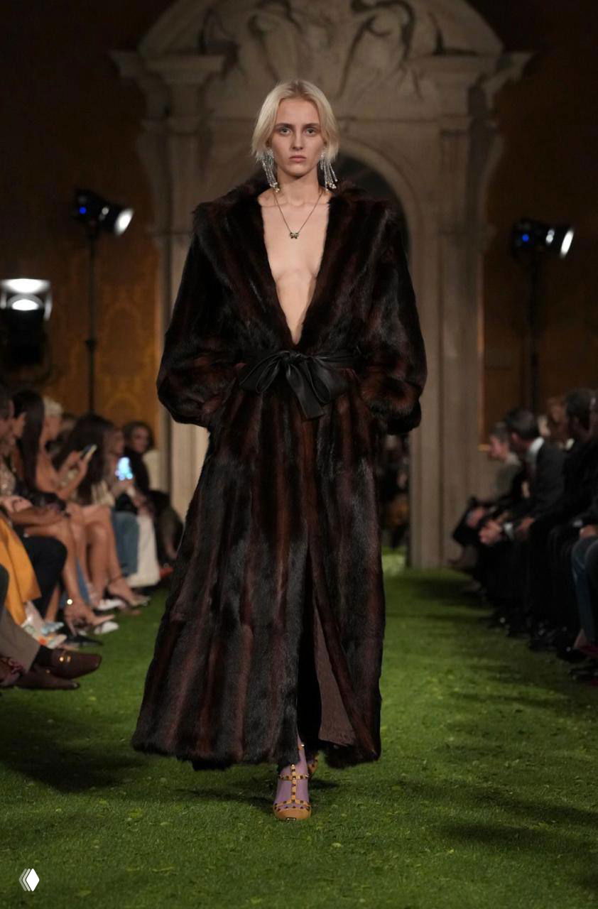 Модель в длинной тёмной шубе с глубоким декольте проходит по зелёному подиуму на показе Valentino в Риме вечером.