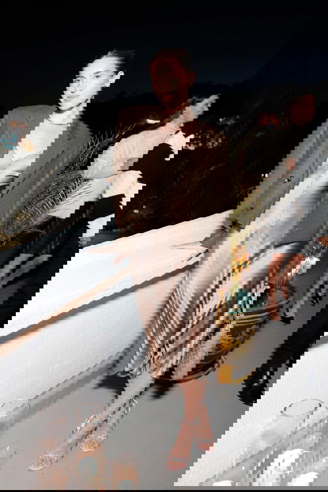 Женщина в бежевой миди-комбинации и укороченном жакете опирается о перила на вечеринке, мягкий блеск ткани подчёркивает силуэт.