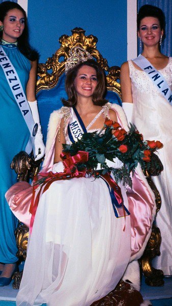 Победительница на тронике с букетом и лентой Miss Universe — портрет в платье, отражающий стандарты красоты и сценический стиль прошлых десятилетий.