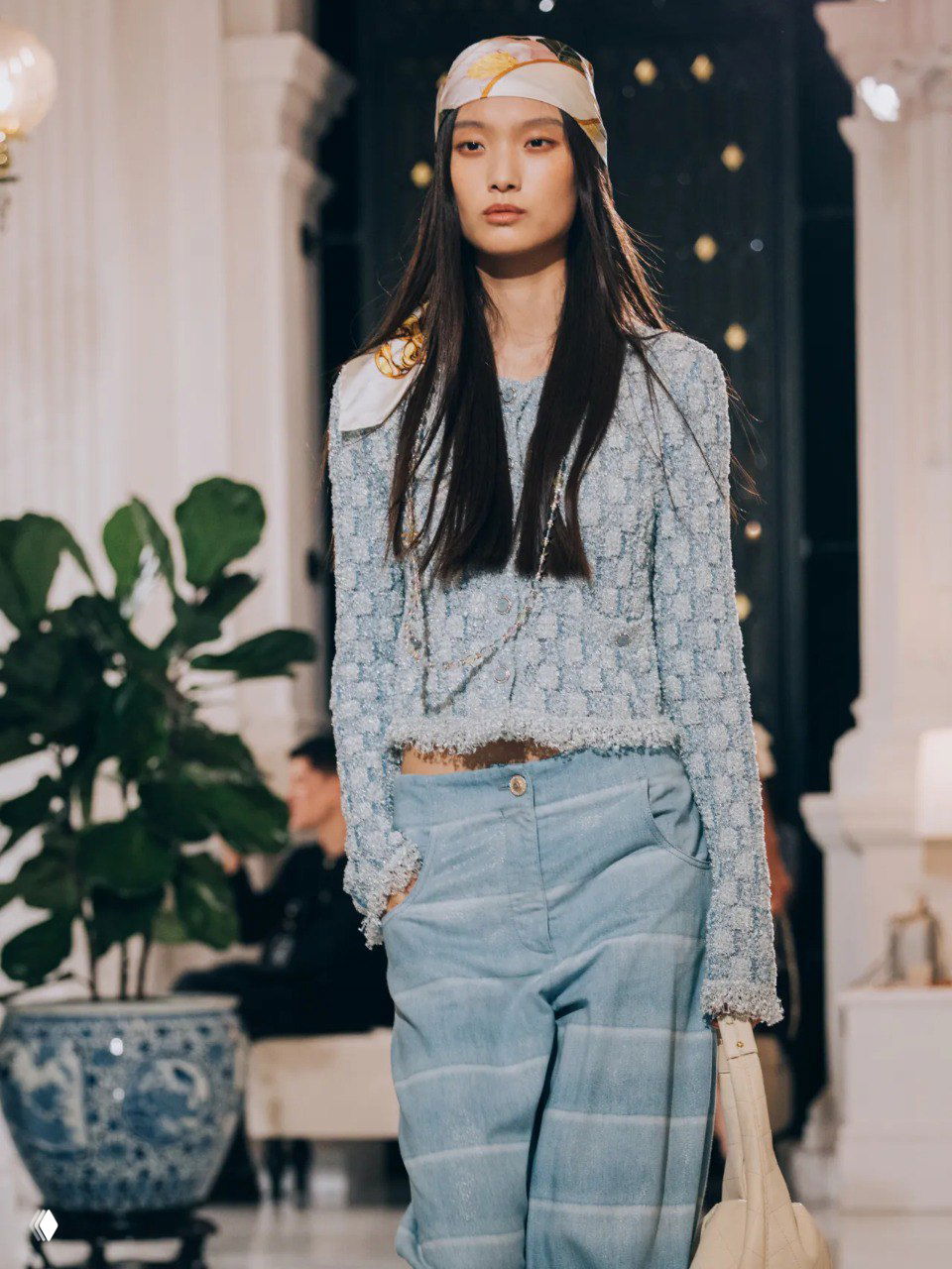 Модель в голубом твидовом топе и свободных брюках с шёлковым платком на голове демонстрирует расслабленный пляжный характер круизной коллекции на показе.
