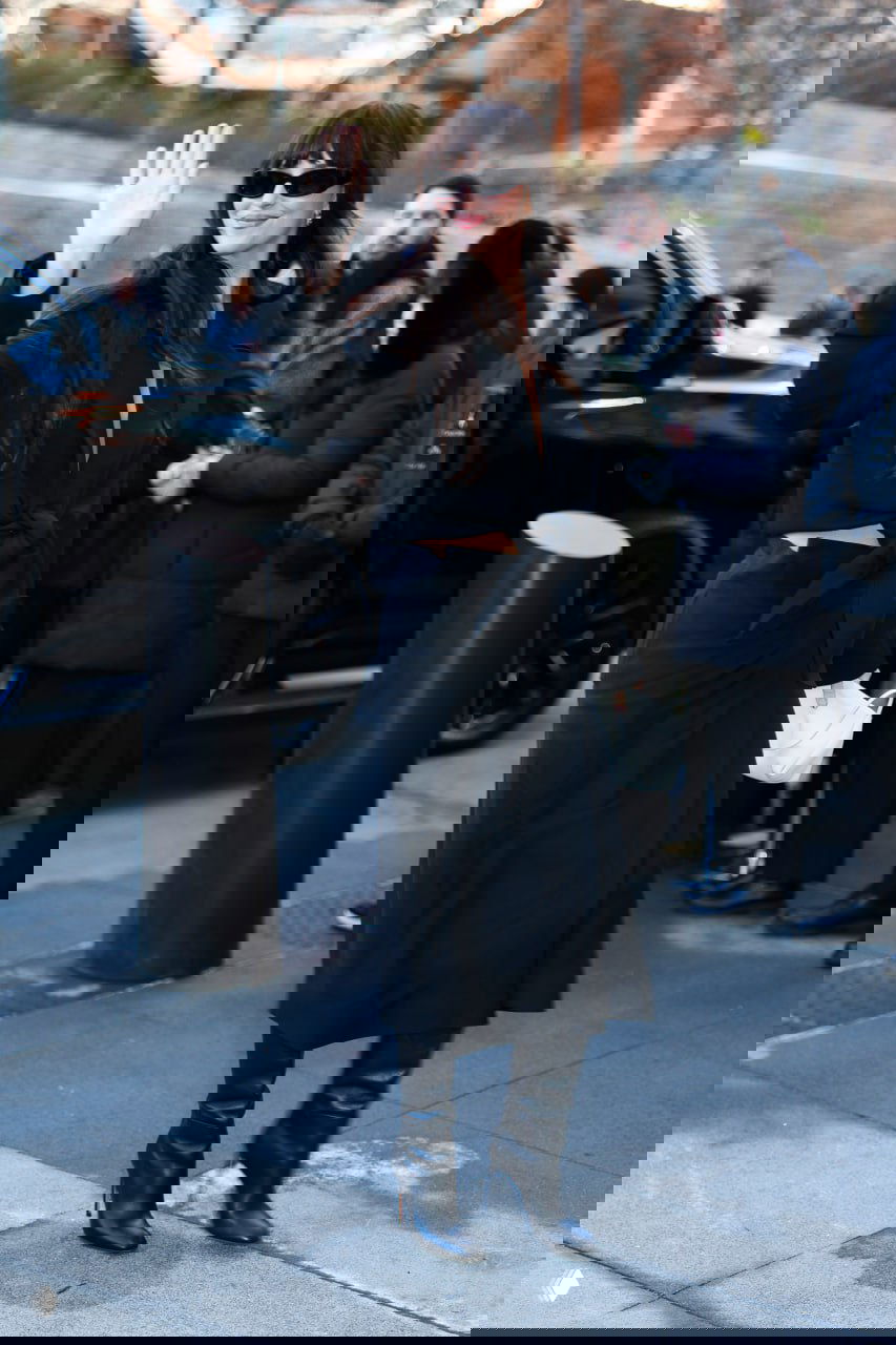 Женщина в полностью чёрном наряде машет рукой; солнцезащитные очки и сапоги создают дерзкий образ на публике.