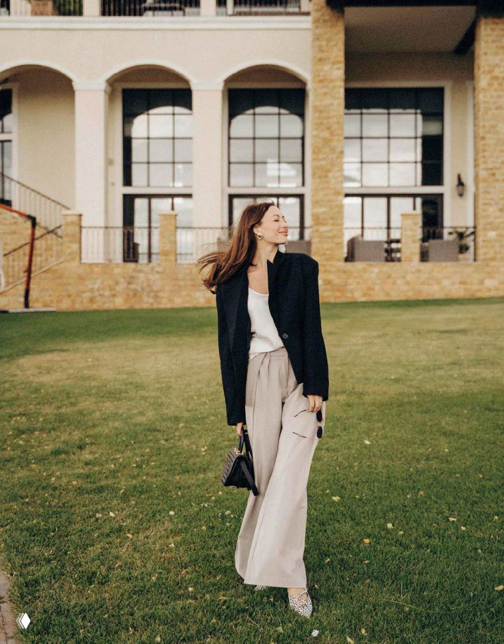 Полноразмерный снимок женщины в повседневном образе: пиджак, широкие брюки и сумка на фоне архитектуры и газона, элегантная уличная съемка.