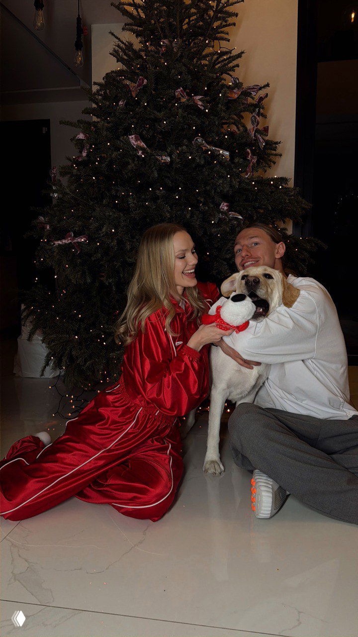 Марина и Матвей играют с лабрадором Морти у большой нарядной ёлки: собака держит игрушку, пара улыбается в тёплой семейной сцене