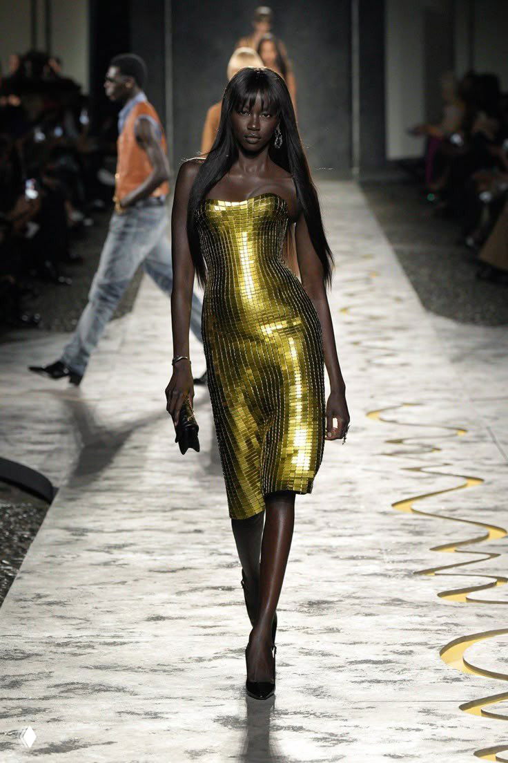 Модель в облегающем золотистом платье идет по подиуму во время показа, на заднем плане — публика и другие модели.