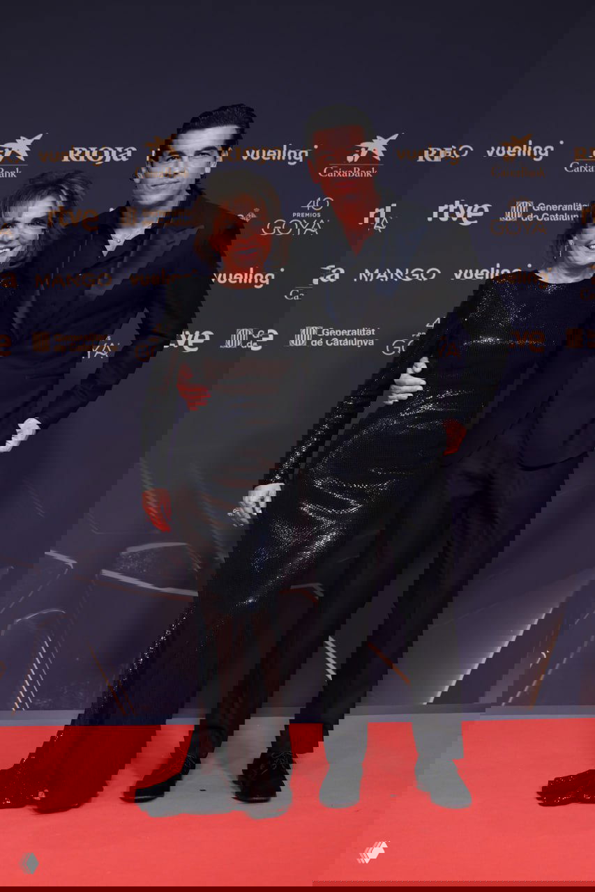 Мужчина в тёмном костюме и женщина в чёрном блестящем платье на красной дорожке церемонии Goya в Барселоне, фон с логотипами.