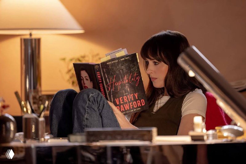 Женщина сидит с книгой в руках: крупный план обложки с названием «Verity Crawford», уютная настольная лампа рядом.