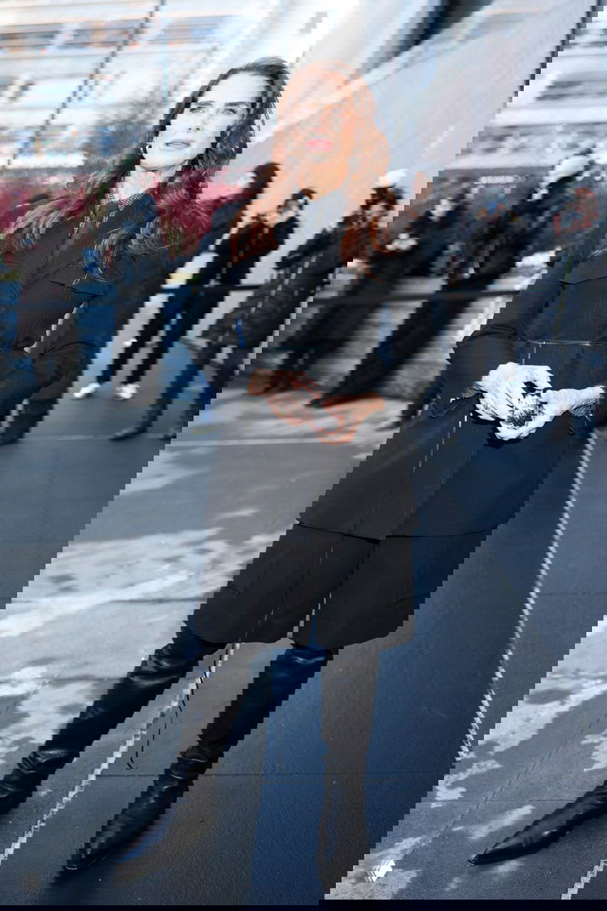 Портретная уличная съёмка: женщина в чёрном костюме и высоких сапогах позирует на фоне зрителей и фотографов.