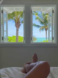 Ноги на кровати у открытого окна с видом на пальмы и бирюзовое море — спокойный уголок с пляжным видом и мягким светом.